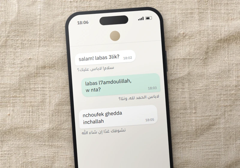 Smartphone posé sur du lin : conversation en darija écrite en arabizi, « salam! labas 3lik? », transcription arabe sous chaque bulle