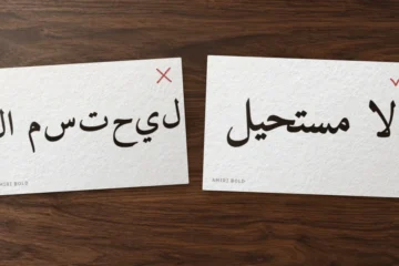 Deux cartes sur un bureau : لا مستحيل en Amiri en lettres isolées posées à l’envers, barré, puis correctement lié, coché